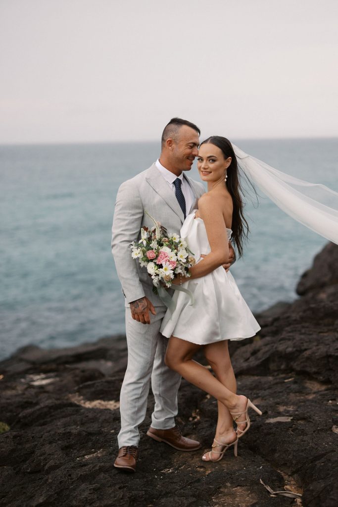 A bride and groom in wedding attire standing on rocks by the ocean, holding a bouquet of flowers, with the groom smiling at the bride and the bride smiling at the camera, with a veil flowing in the wind.