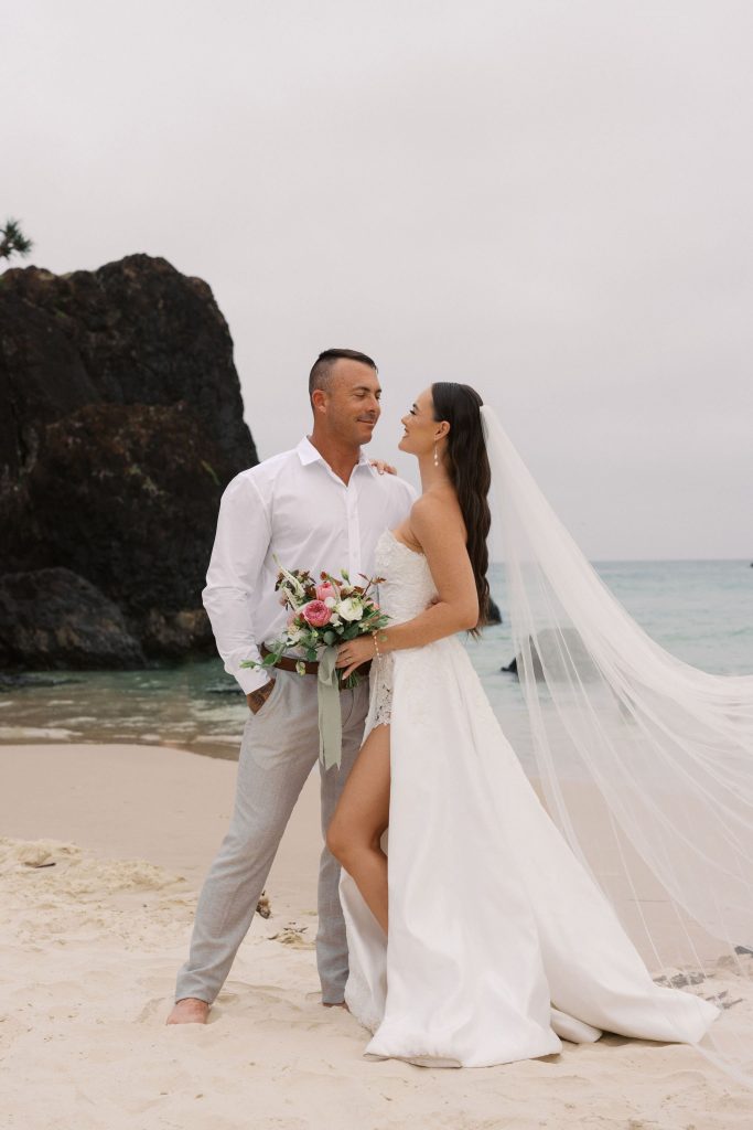A bride and groom embracing. The bride is holding a romantic and textured bouquet design by Hart & Flowers combining the delicate beauty of garden roses, sweetpeas, phlox, and veronica, with soft trailing ribbon image captured on Fingal Head Beach.