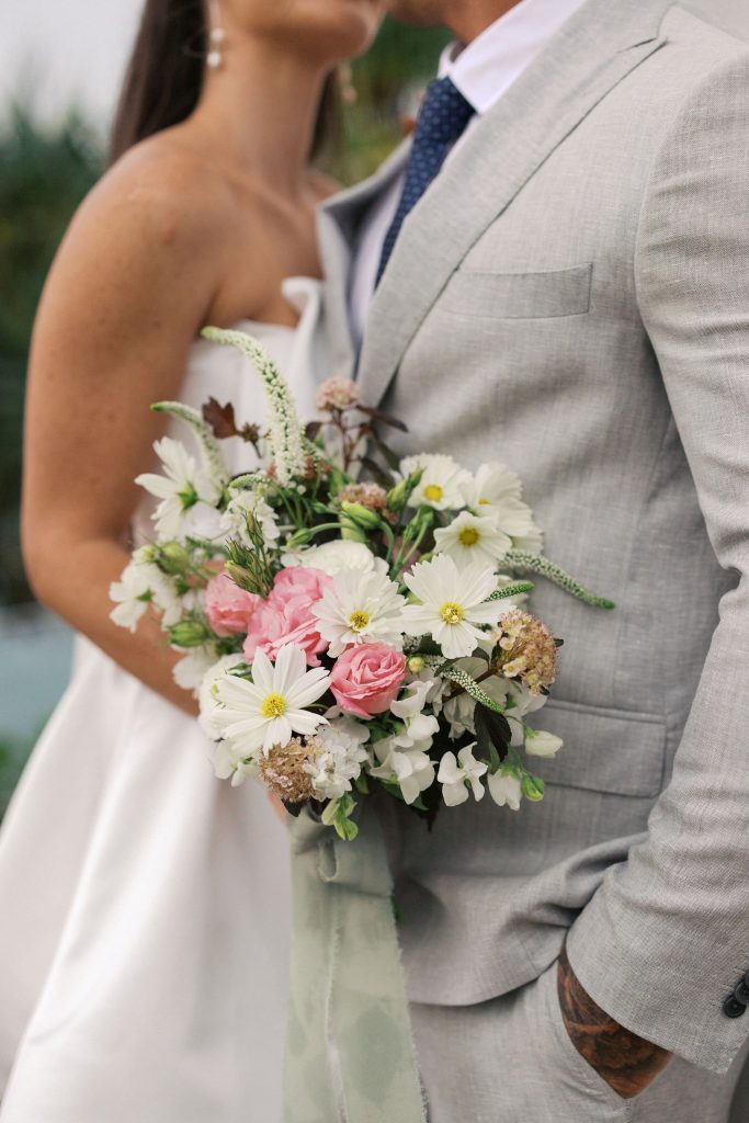 A bride and groom standing close together, with the bride holding a bouquet of pink and white flowers. The groom is dressed in a light gray suit. The bride's face is not visible, and they are outdoors.