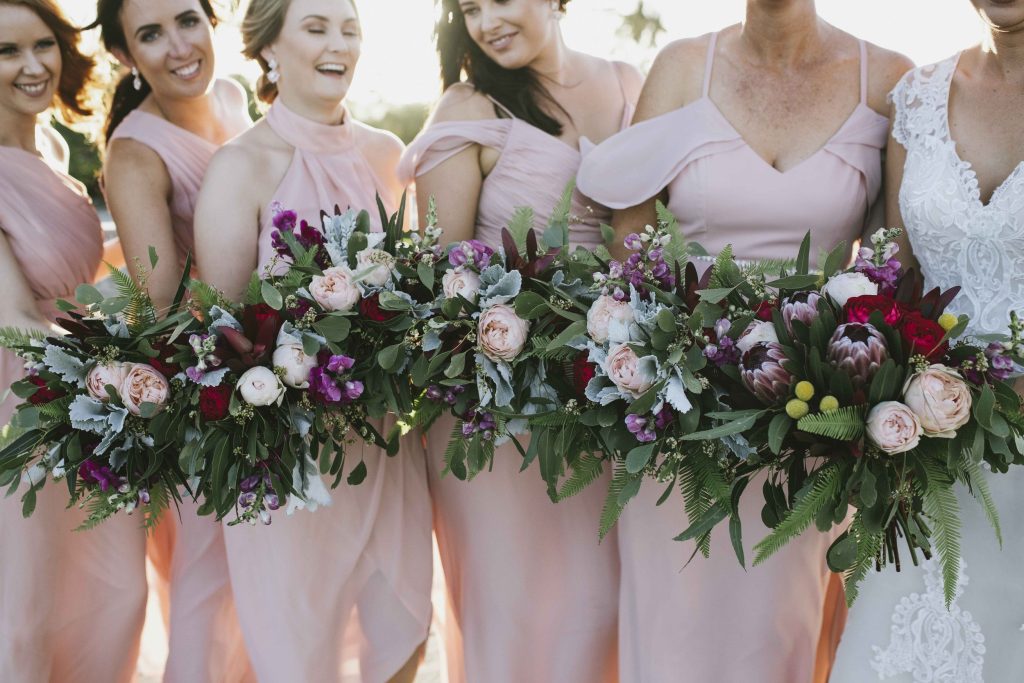 Group of women in pink dresses holding a large floral arrangement with pink, white, purple, and red flowers along with green foliage.