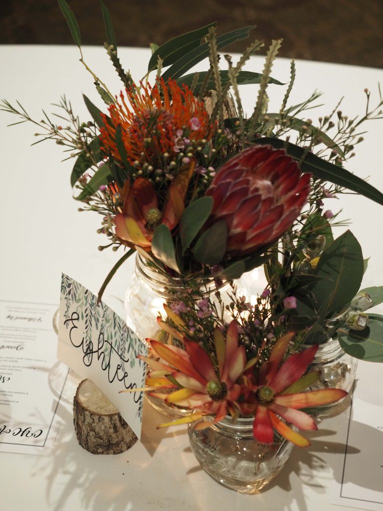 Two small glass jars filled with various colorful flowers on a white table, with a small wood slice and a sign nearby.