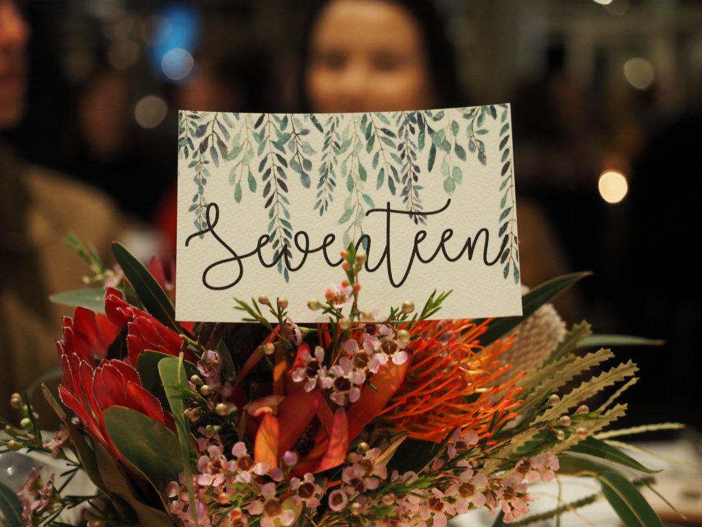 A floral arrangement with a card that says "Seventeen" on it, featuring pink and red flowers, green leaves, and rustic decor, with a blurred background of people.