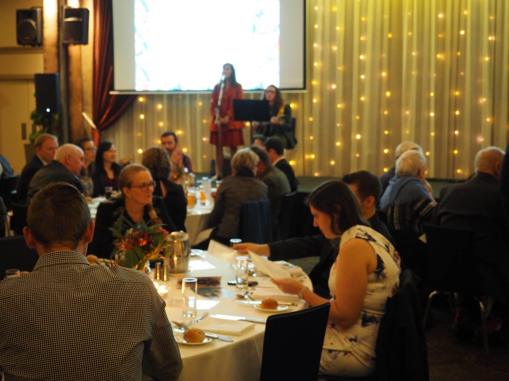 People attending a formal event or banquet, sitting at round tables with white tablecloths, in a decorated hall with string lights, with a woman at a podium speaking and a man playing keyboard on stage in the background.