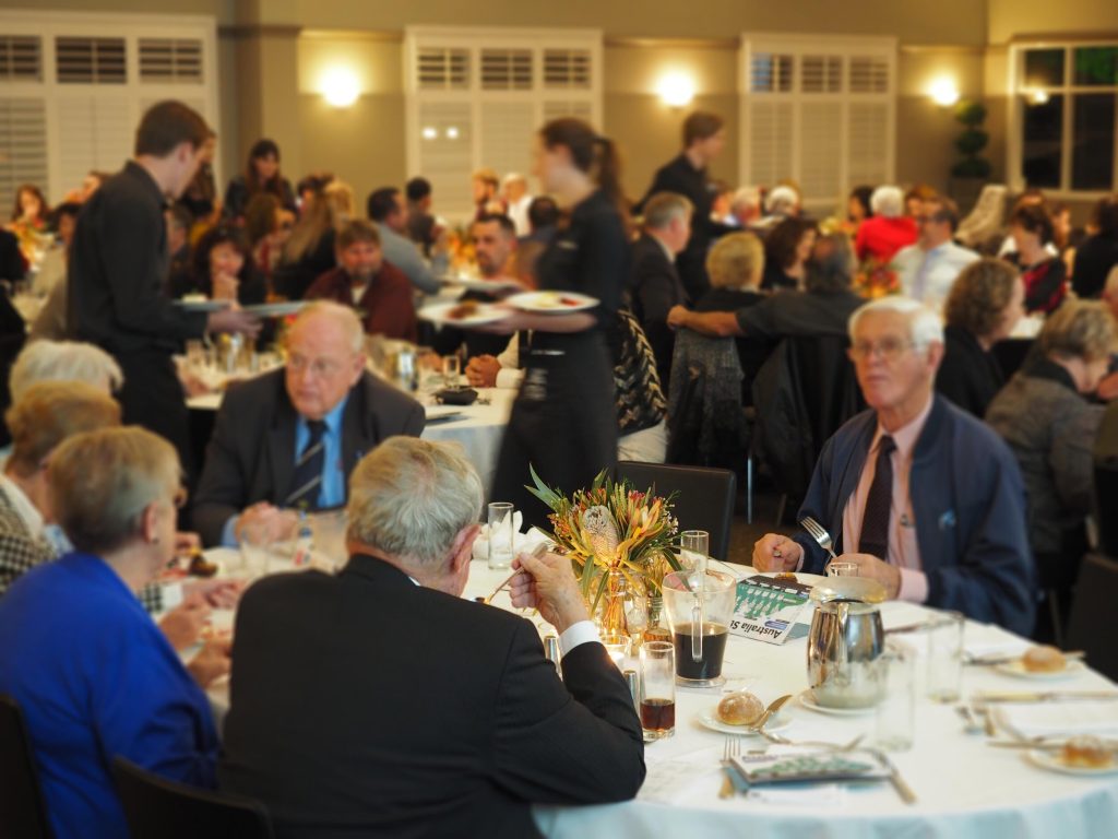 A busy banquet hall with many people dining at round tables, waitstaff serving food, and a large floral centerpiece on one table.