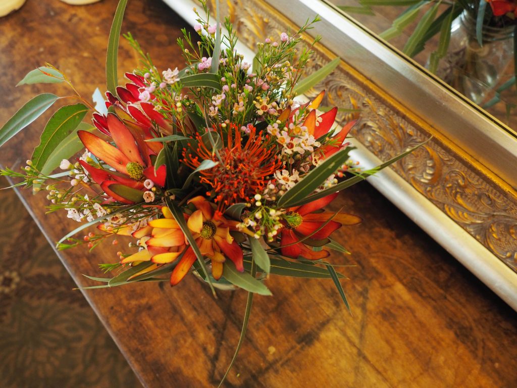 Colorful flower arrangement featuring red, orange, and yellow flowers with green leaves on a wooden surface, near a framed mirror with ornate gold detailing.