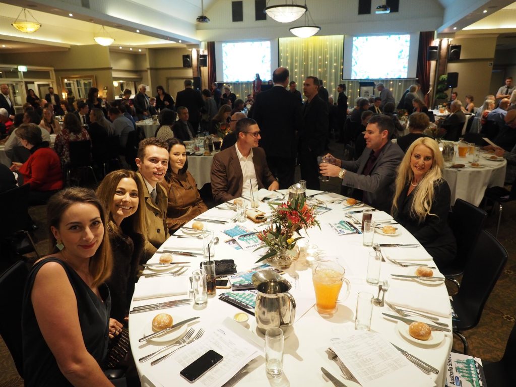 A group of seven people seated at a round banquet table during a formal event in a large, well-lit room, with many other attendees in the background. The table is set with plates, glasses, cutlery, and a centerpiece of flowers. Some people are holdin