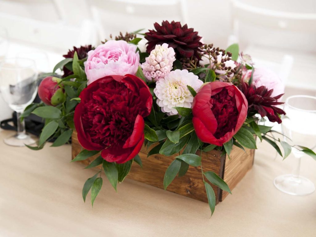 A vibrant and romantic wedding table setting featuring a centerpiece of deep red and pink blooms arranged in a rustic wooden box. The arrangement showcases a mix of lush peonies and dahlias, creating a bold and eye-catching display. The wooden box ad