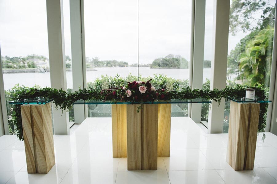 A modern and elegant wedding ceremony setup with a glass table adorned with a cascading garland of greenery and vibrant blooms. The centerpiece features a mix of deep red and pink flowers, adding a touch of romance and drama. The breathtaking view of