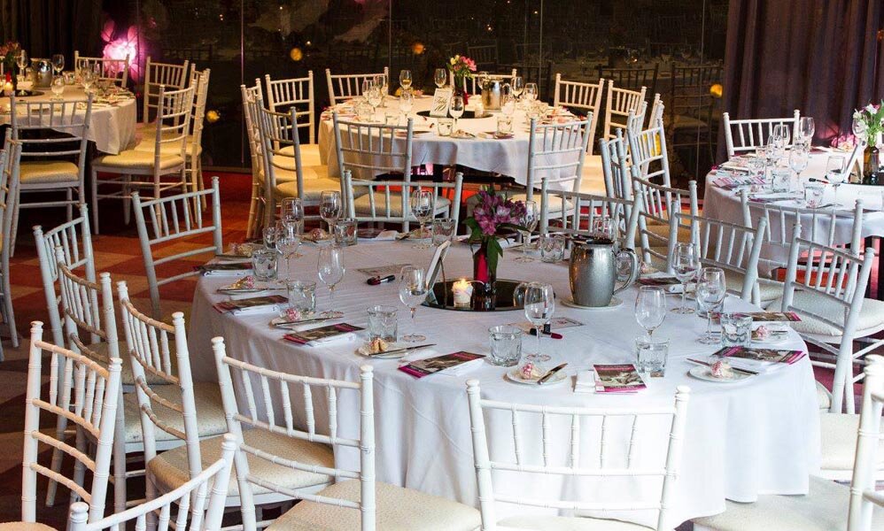 Elegant banquet hall with round tables covered in white tablecloths, set with glasses and silverware, surrounded by white chairs, decorated with floral centerpieces.