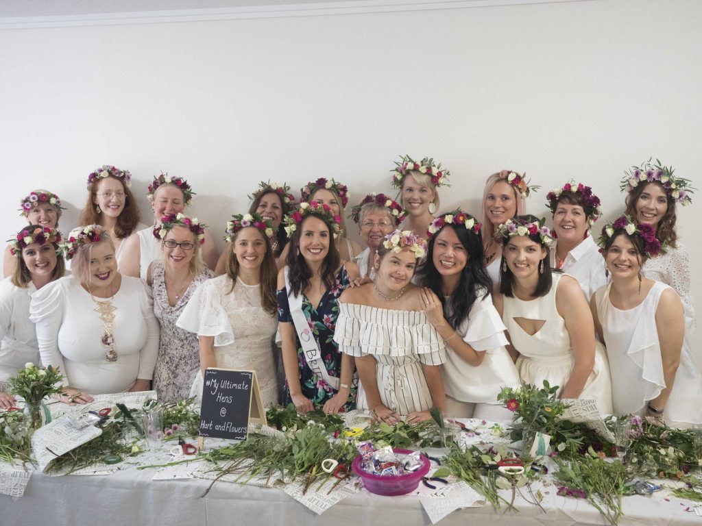 A group of women and young girl celebrating a special occasion, all wearing white dresses and flower crowns, in front of a decorated table with flowers and craft supplies.