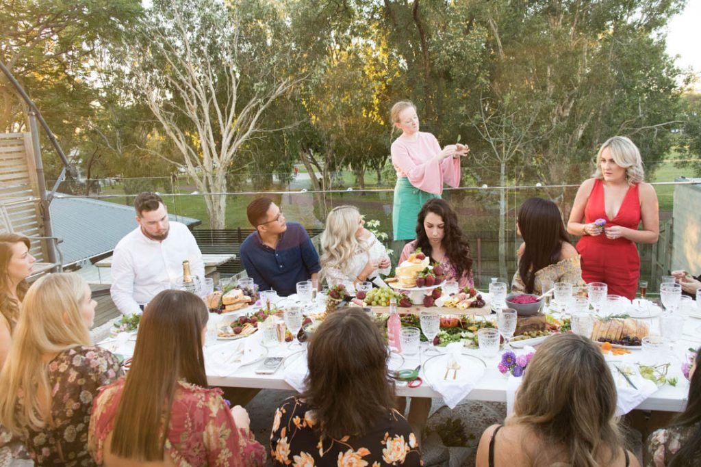 A group of people enjoying a dinner party outdoors with a long table filled with food and drinks, surrounded by trees and greenery.