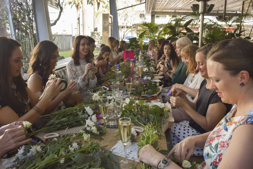 Women participating in a flower arranging workshop at a long table outdoors, surrounded by flower arrangements, scissors, and framed by tropical plants.