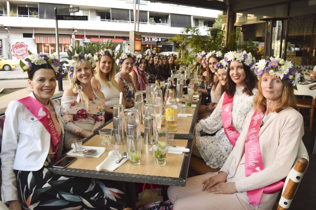 A group of women wearing flower crowns and pink sashes sitting at a long table celebrating a bridal shower, with drinks and party supplies on the table.