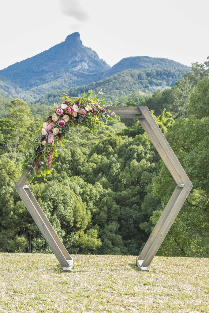 A natural and elegant floral design by Hart & Flowers graces a hexagonal arbour at Mavis's Kitchen Weddings, showcasing protea, banksia, garden roses, trailing burgundy amaranthus and flowing greenery creates a beautiful ceremony backdrop with breath