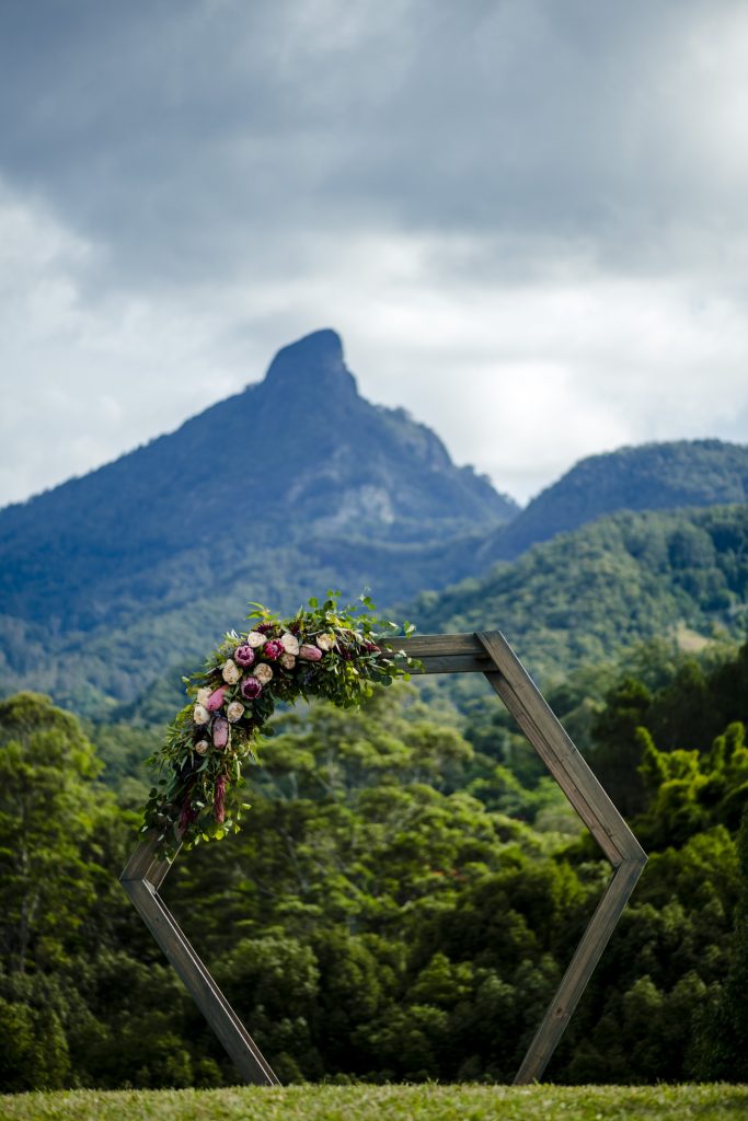 A wooden hexagonal frame decorated with flowers, set outdoors in front of a lush green forest and a mountain with a distinctive peak under a cloudy sky.