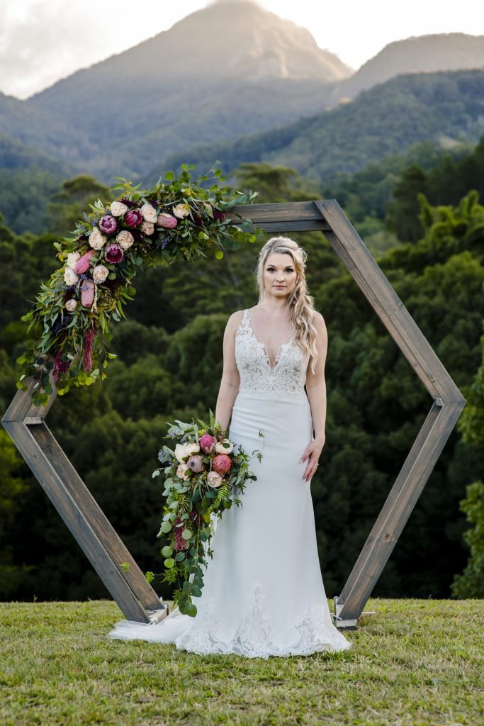 A stunning wedding ceremony setup with a rustic wooden arch adorned with a cascading arrangement of pink and red flowers. The arch is set against a breathtaking backdrop of the majestic Wollumbin National Park, creating a dramatic and unforgettable s
