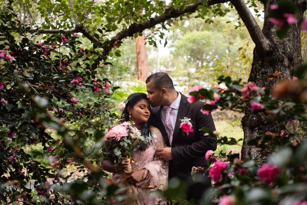 A couple dressed in wedding attire sharing a tender moment in a garden with pink and purple flowers and green foliage.