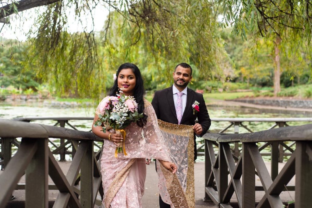 A couple dressed in traditional attire standing on a wooden bridge near a pond, holding a bouquet of flowers, with trees and greenery in the background.
