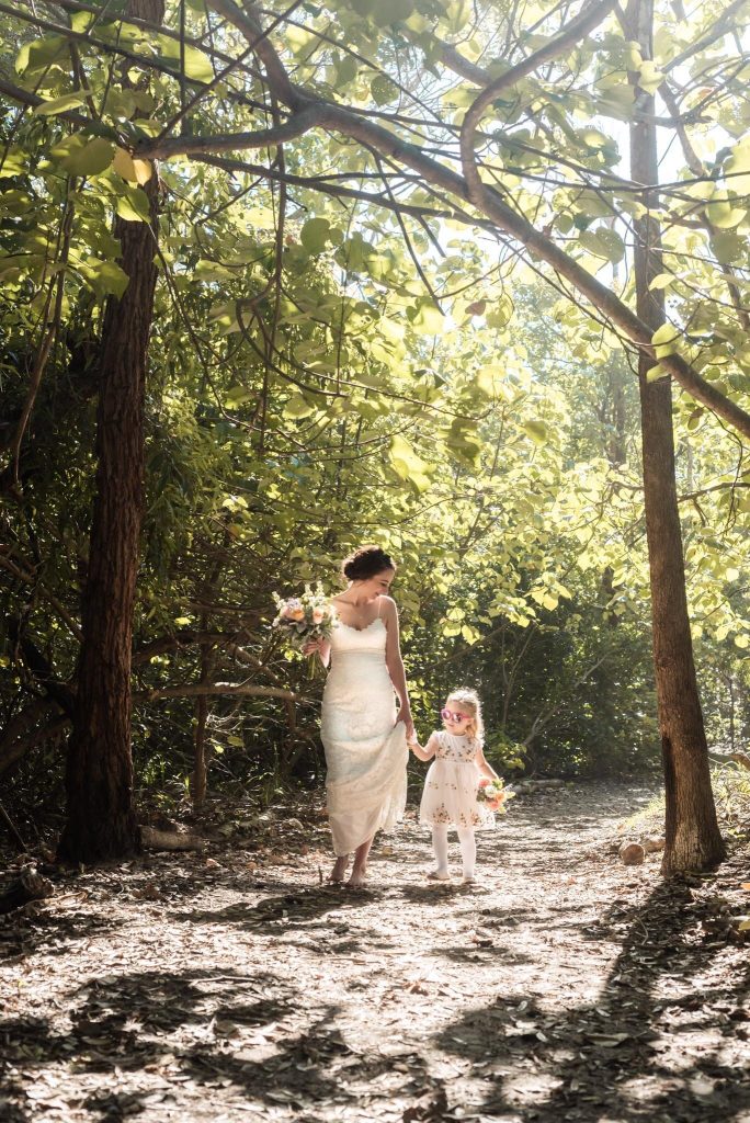 A woman in a white wedding dress holding a bouquet and a young girl in a floral dress with pink sunglasses walking hand-in-hand through a wooded area.