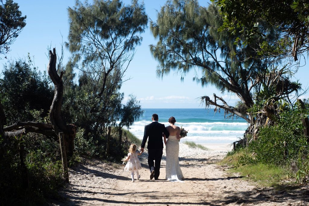 A couple in wedding attire walking hand in hand with a young girl on a sandy beach pathway, heading towards the ocean, framed by trees and blue sky.