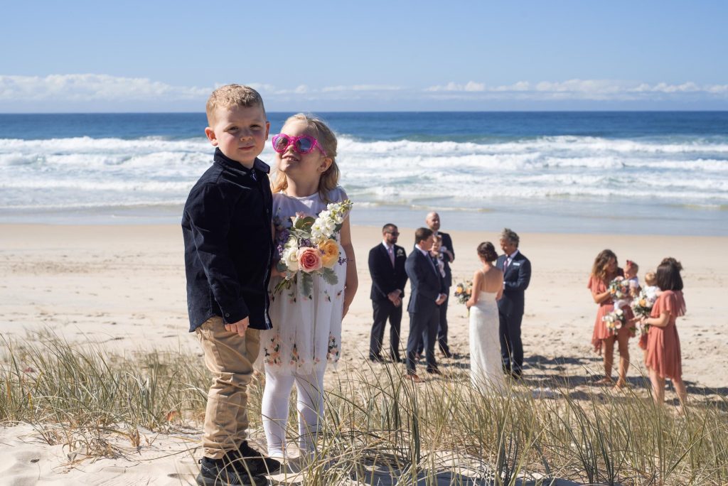 A wedding ceremony on the beach with children in the foreground and the bride, groom, and guests in the background.