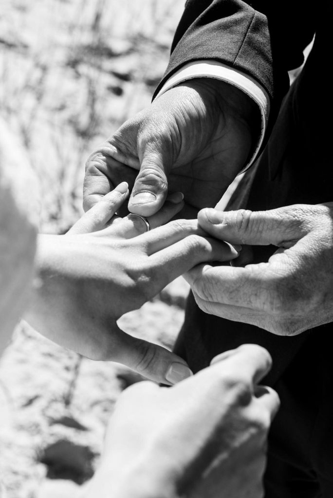 Close-up of two people's hands holding a wedding ring, with a third person's hand passing it to one of them, outdoors, in black and white.