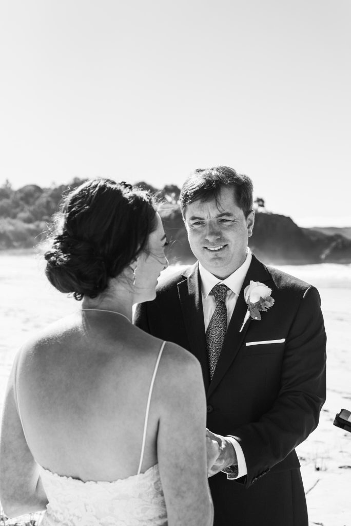 A black and white photograph of a couple during a wedding ceremony on the beach, with the woman in a lace dress and the man in a suit with a boutonniere, holding hands.