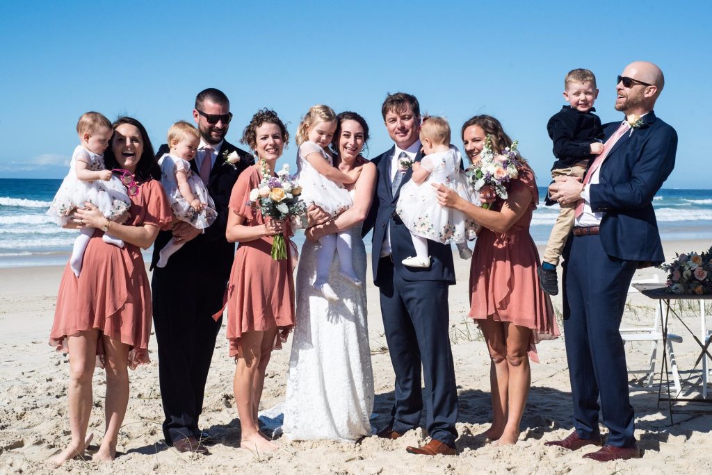 Group of people at a beach wedding, including bride and groom, bridesmaids, groomsmen, and children, with the ocean in the background.