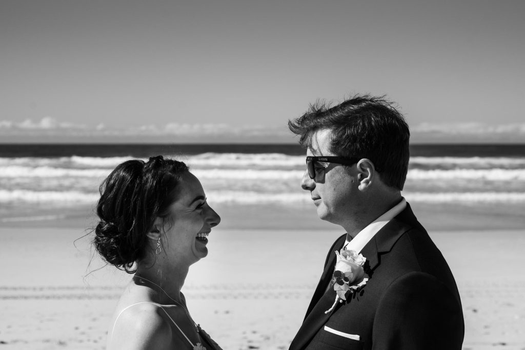 A black and white photograph of a bride and groom standing face to face on a beach, smiling at each other. The bride has dark hair styled in an updo and is wearing a sleeveless dress. The groom is wearing sunglasses, a suit with a boutonniere, and ha