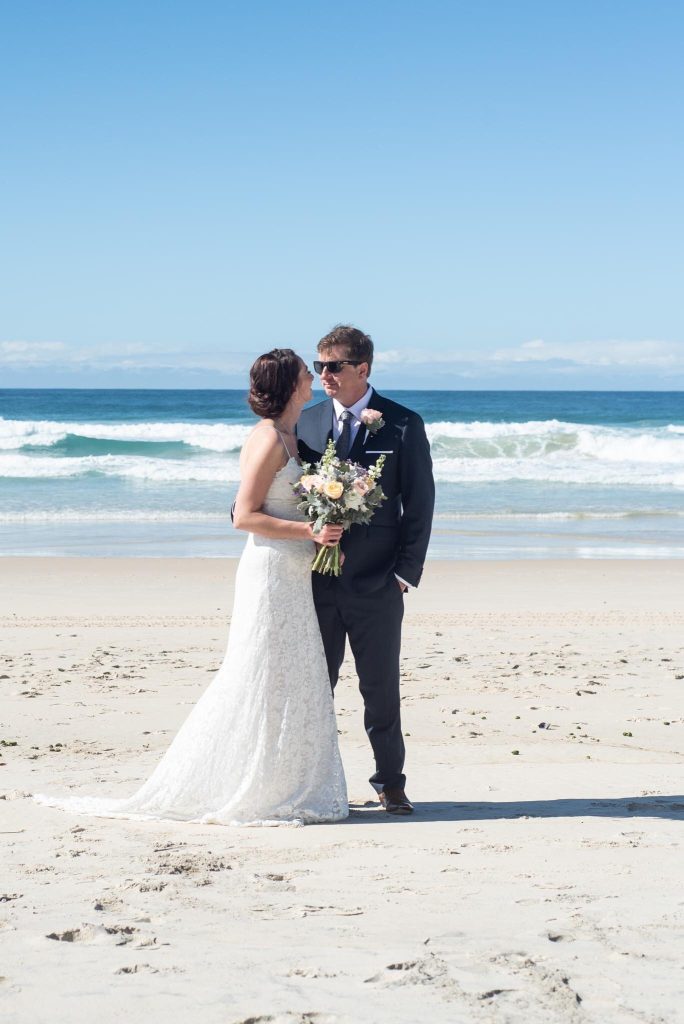 A bride and groom standing on a sandy beach with ocean waves in the background, during a wedding photoshoot.