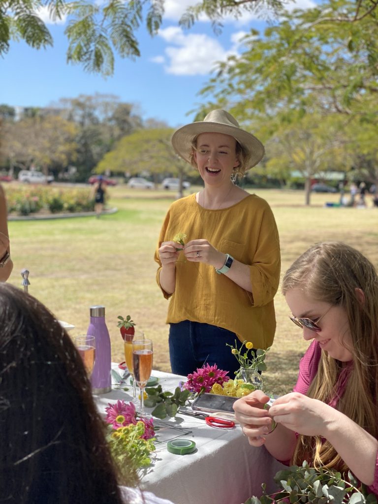 A woman in a yellow top and wide-brimmed hat standing outdoors at a table with flowers, champagne flutes, and gardening supplies. Partially visible women are sitting at the table, engaging in flower arranging or crafting activities, in a park with tr