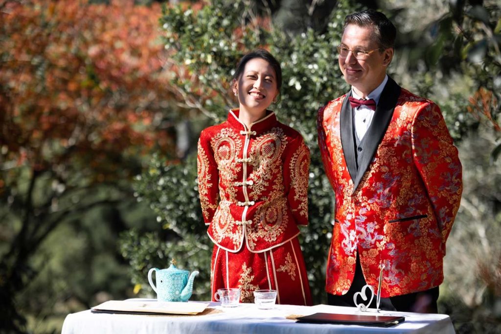 A woman and a man dressed in traditional Chinese and formal tuxedo wedding attire, smiling outdoors with a table set with a teapot and cups in front of them.