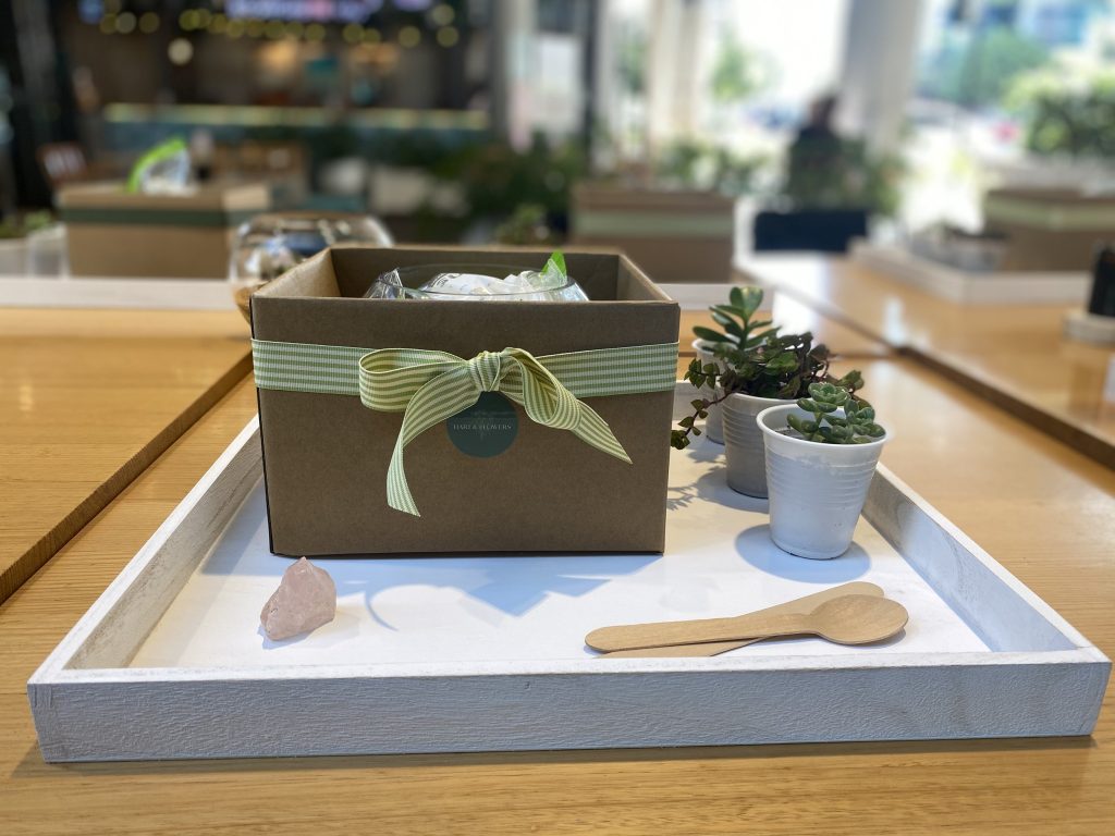 Decorative tray with potted succulents, a small wooden spoon, a pink rock, and a gift box with a green and white ribbon inside a cafe or restaurant setting.