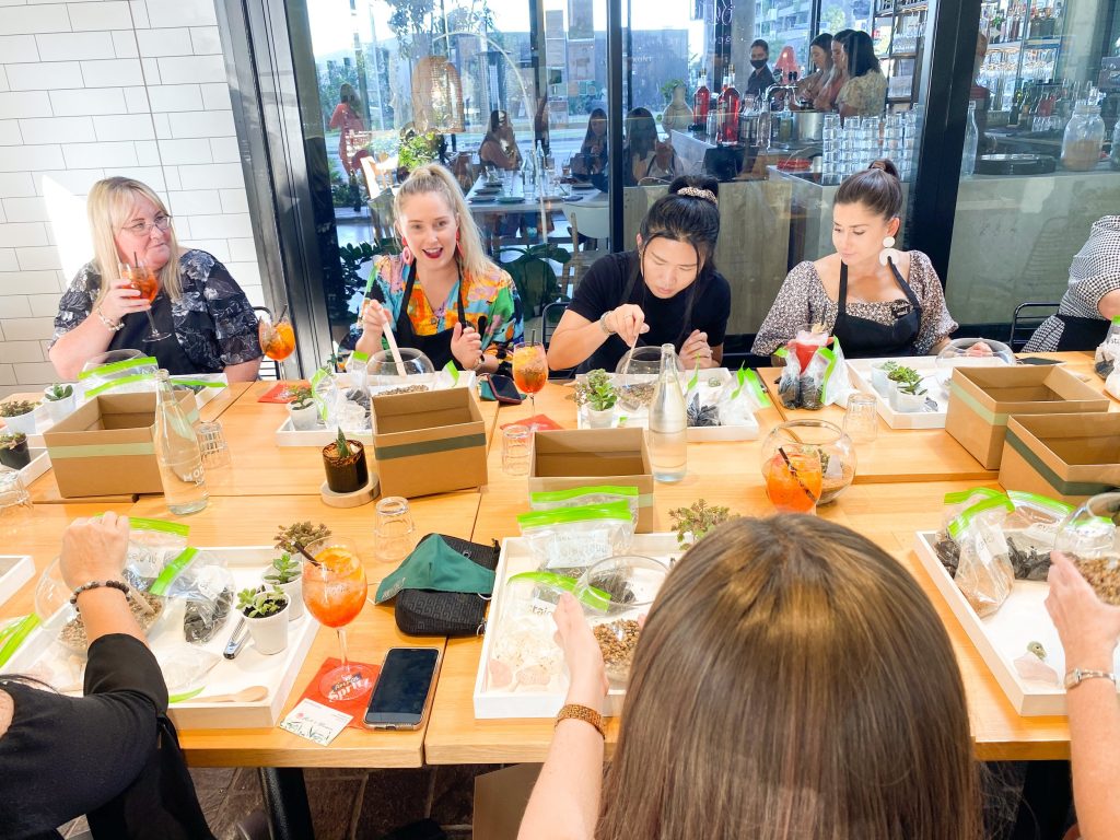 Group of women sitting around a wooden table with catering boxes and drinks, participating in a planting activity at a restaurant.