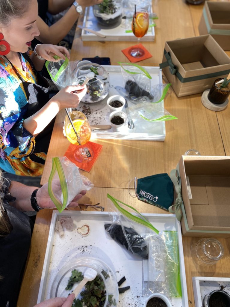 People participating in a plant terrarium workshop, handling soil, plants, and decorative items at a wooden table.