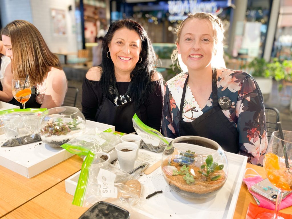 Two women sitting at a table with desserts and plants, smiling at the camera, in a brightly lit indoor setting.