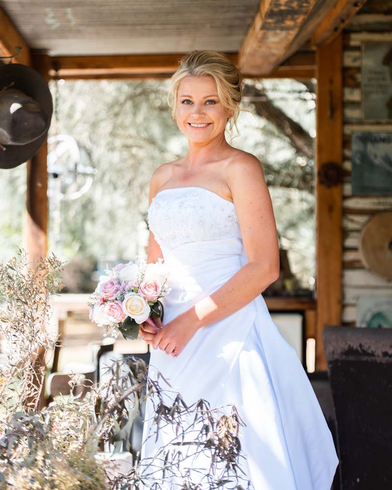 A woman in a strapless white wedding dress holding a bouquet of pink and white roses inside a rustic wooden structure with greenery outside.