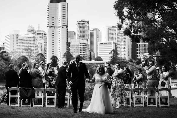 A bride and groom walking down the aisle holding hands at an outdoor wedding ceremony in a park with city skyscrapers in the background.