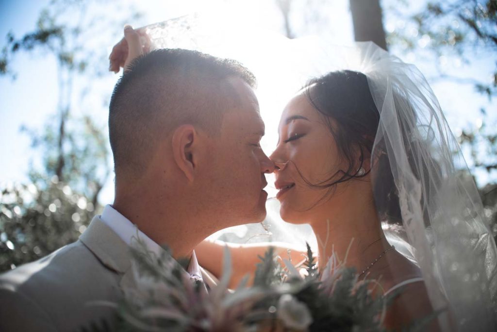 A bride and groom sharing a kiss outdoors with sun shining behind them.