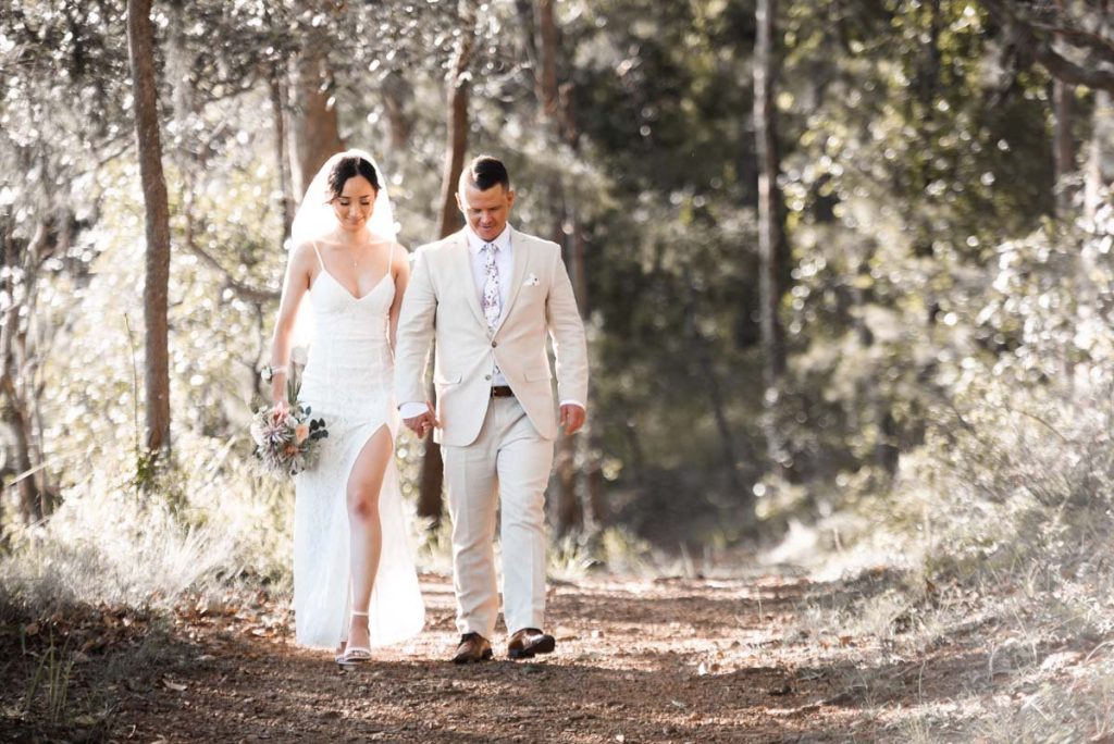 A bride and groom walking together on a dirt path through a wooded area during their wedding, holding hands and smiling.
