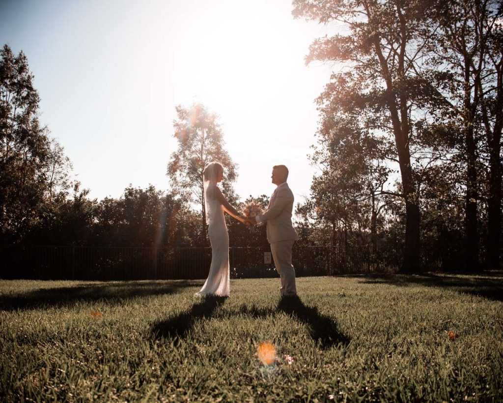 A bride and groom holding hands and facing each other outdoors during sunset, with trees in the background and shadows on the grass.