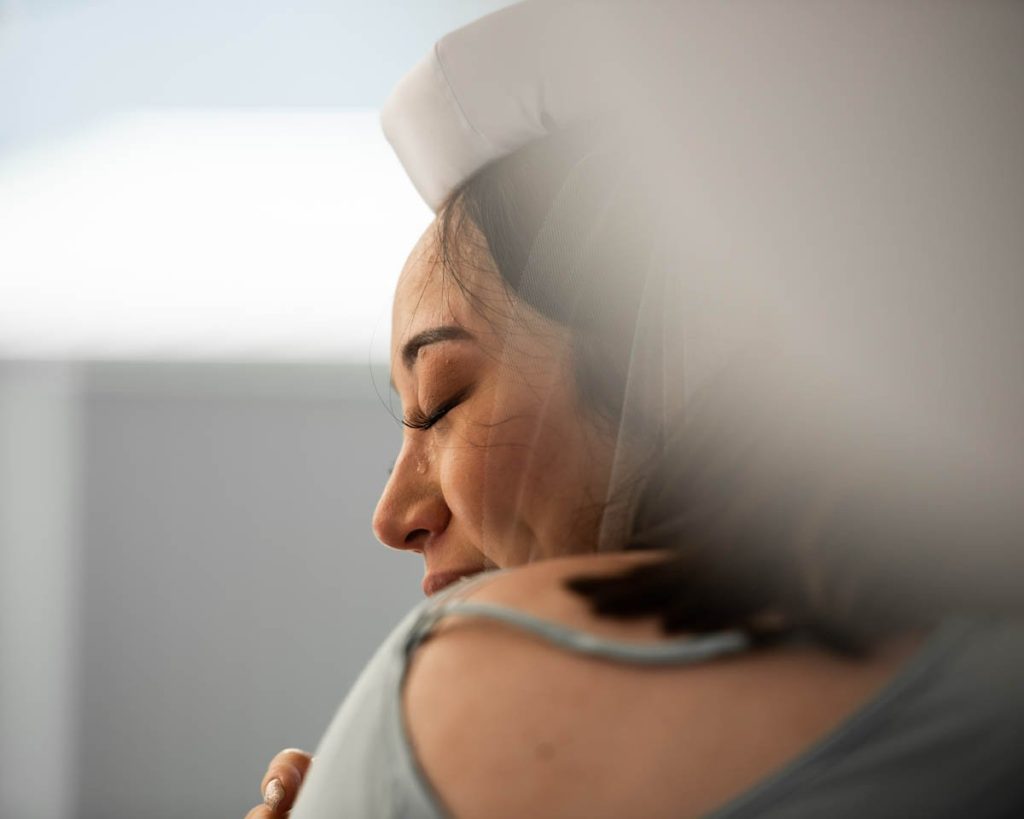 A woman with a tear falling down her face, her eyes closed, hugging her shoulder, with a nurse or healthcare worker in a white uniform and cap gently comforting her.