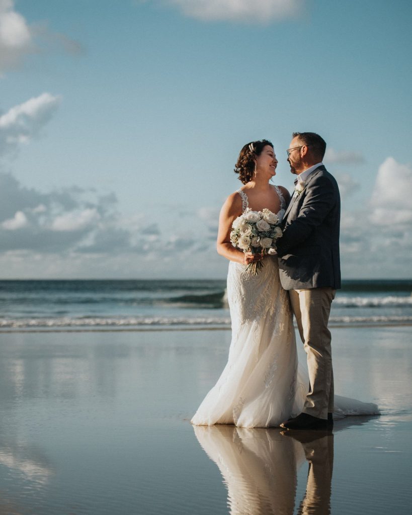 A bride and groom standing together on a beach during their wedding, holding a bouquet of flowers and looking happily at each other.