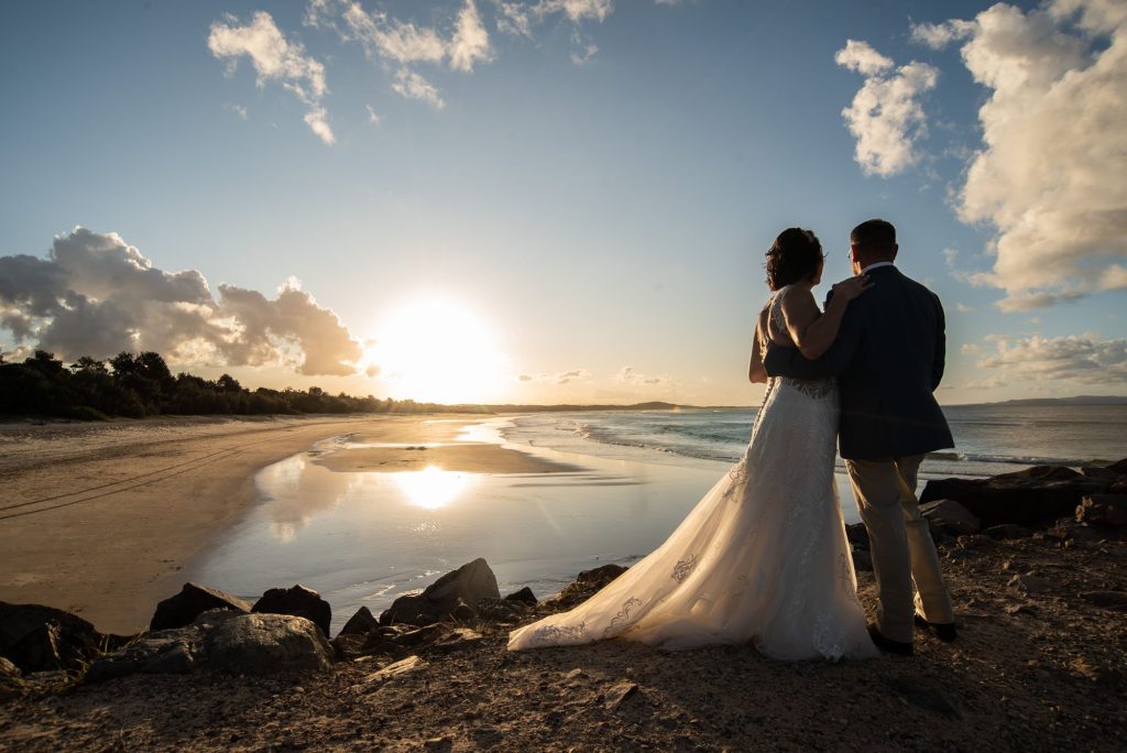 A bride and groom stand close together on a rocky beach at sunset, holding each other and gazing at the horizon with the ocean and sky in the background.