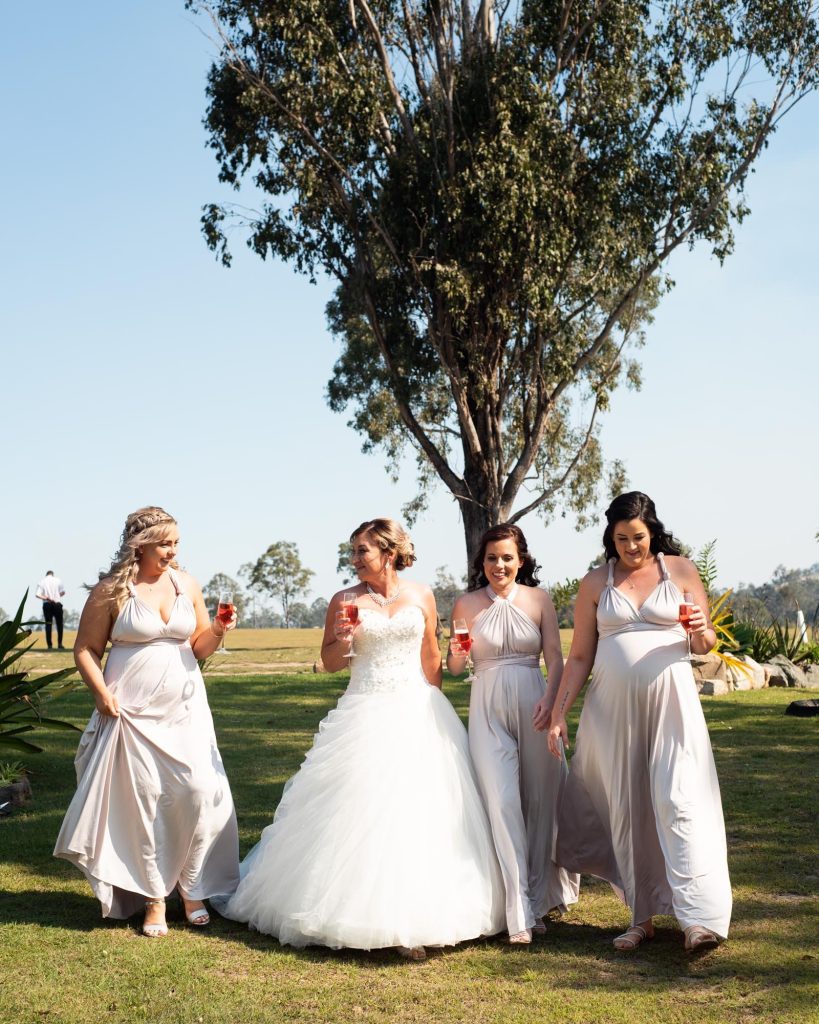 Bride in white wedding gown walking with four bridesmaids in gray dresses outdoors on a sunny day, each holding a glass of pink drink, near a large tree.