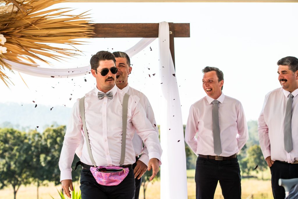A group of men in white shirts and gray ties celebrating at an outdoor wedding, with one man wearing sunglasses and a pink fanny pack, as confetti falls around them.