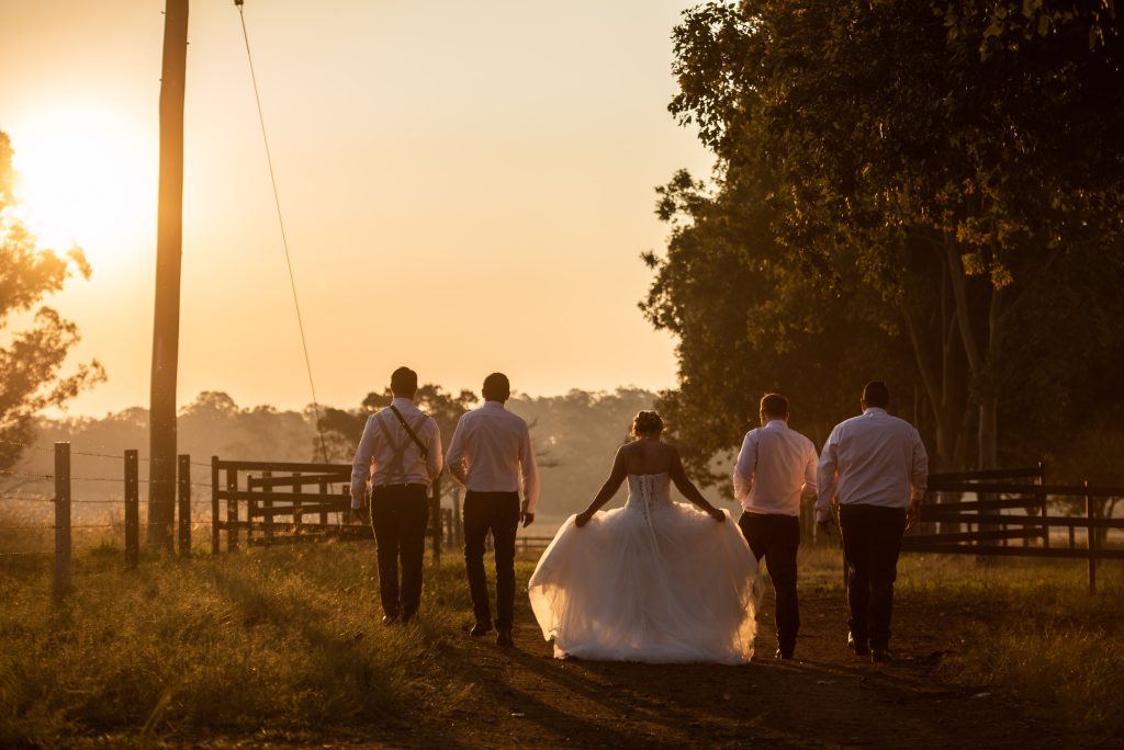 A bride in a white wedding dress walking with four men in formal attire on a dirt road at sunset.