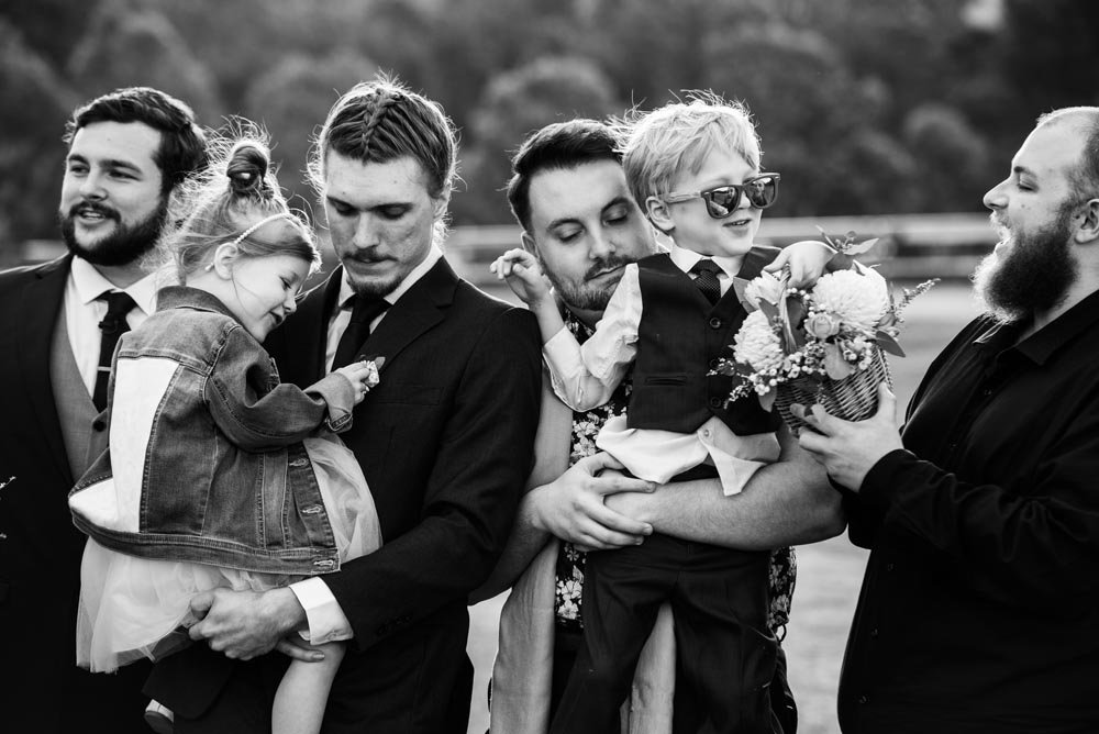 Black and white photo of a group of men and children gathering outdoors, with one man handing a bouquet of flowers to a young boy wearing sunglasses.