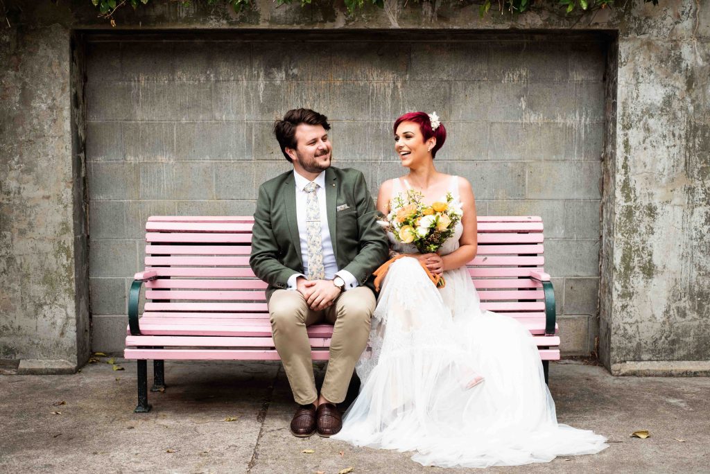 A happy bride and groom sitting on a pink park bench, sharing a moment of joy. The bride is holding a bouquet of yellow and white flowers, wearing a white wedding dress, and has short red hair. The groom is dressed in a green suit jacket, a patterned