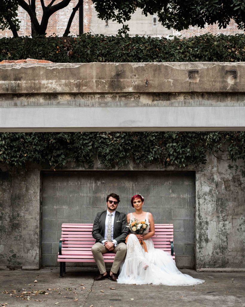 A bride and groom sitting on a pink park bench under a concrete archway with ivy and trees in the background.